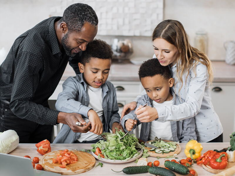 Mom And Dad Sharing Salad Recipe, Multiracial Family Chatting Enjoying Time In Kitchen.